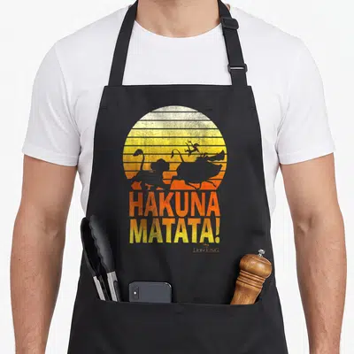 Tablier de cuisine noir avec silhouettes de personnages du Lion King devant un soleil orange et le texte Hakuna Matata.