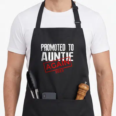 Tablier de cuisine noir avec linscription « Promoted to Auntie Again 2023 » en blanc et rouge.