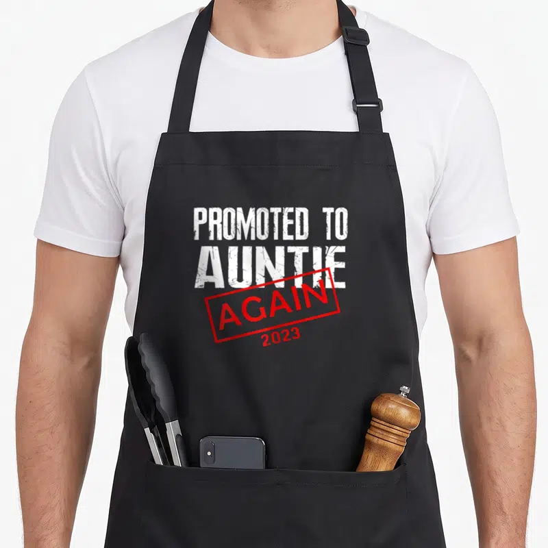 Tablier de cuisine noir avec linscription « Promoted to Auntie Again 2023 » en blanc et rouge.