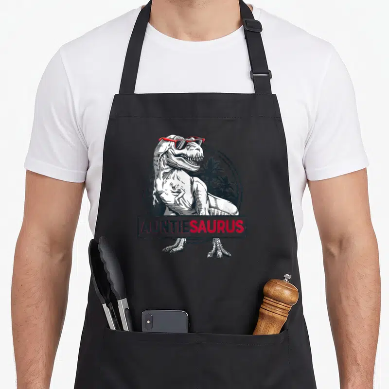 Tablier de cuisine noir avec illustration d'un T rex portant des lunettes rouges et le texte AUNTIESAURUS en bas.