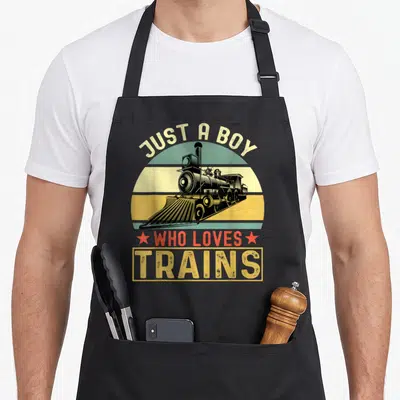 Tablier de cuisine noir avec illustration vintage d'une locomotive et le texte Just a boy who loves trains en lettres jaunes et rouges.