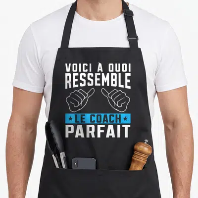 Tablier de cuisine noir avec inscription voici à quoi ressemble le coach parfait et deux mains pointant vers le texte central.