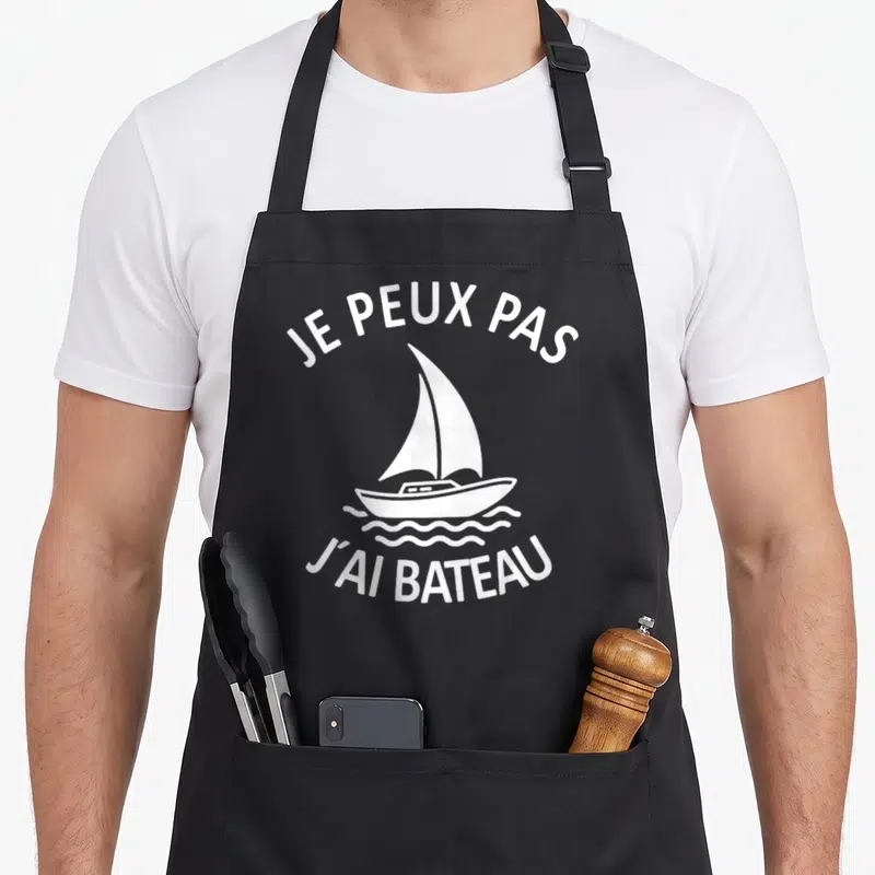 Tablier de cuisine noir avec inscription Je peux pas j'ai bateau et illustration d'un voilier blanc sur fond uni.