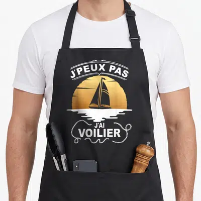 Tablier de cuisine noir avec illustration d'un voilier au coucher du soleil et texte blanc Je peux pas j'ai voilier.