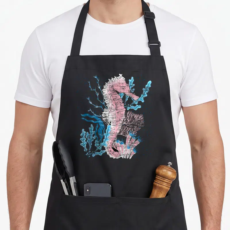 Tablier de cuisine noir avec illustration colorée d'un hippocampe rose et de végétaux marins bleus sur fond pointillé.