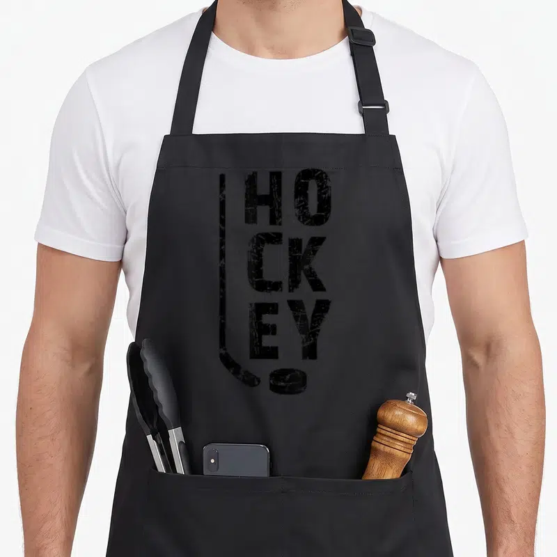 Tablier de cuisine noir avec typographie verticale HOCKEY, crosse de hockey et palet en style usé.