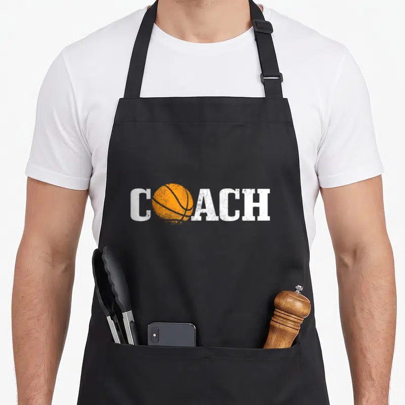 Tablier de cuisine noir avec le mot coach et une balle de basket orange stylisée en centre du design imprimé.