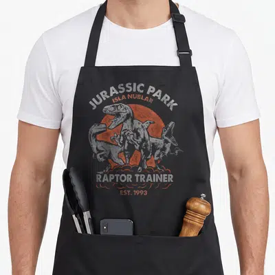 Tablier de cuisine noir avec illustration de raptors et texte Jurassic Park Raptor Trainer Isla Nublar Est 1993 en blanc et orange.