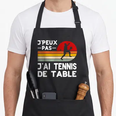 Tablier de cuisine noir avec motif rétro « J'peux pas j'ai tennis de table » et silhouette de joueur sur fond de soleil coloré.
