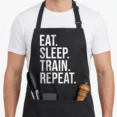 Tablier de cuisine noir avec le texte blanc vertical Eat Sleep Train Repeat imprimé au centre du torse.