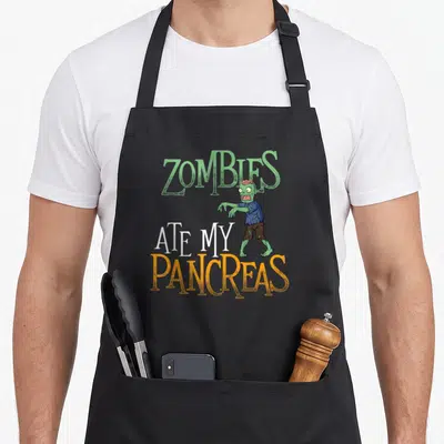 Tablier de cuisine noir avec un zombie vert et le texte Zombies Ate My Pancreas en lettres colorées.