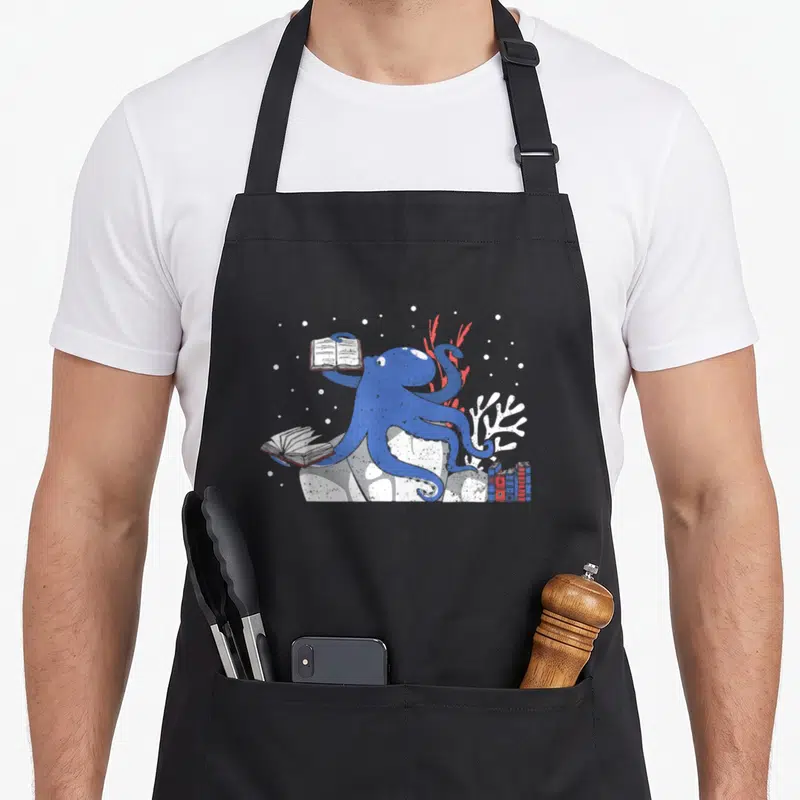 Tablier de cuisine noir avec un poulpe bleu tenant un livre ouvert au milieu de coraux et d'ouvrages posés.