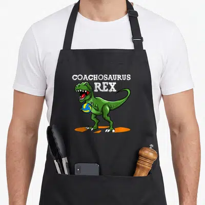 Tablier de cuisine noir avec un dinosaure vert tenant un ballon de volley et le texte Coachosaurus Rex en blanc.