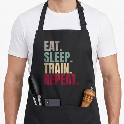 Tablier de cuisine noir avec le texte Eat Sleep Train Repeat imprimé en lettres colorées vertes, jaunes et rouges.