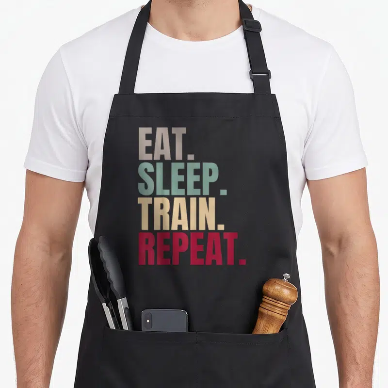 Tablier de cuisine noir avec le texte Eat Sleep Train Repeat imprimé en lettres colorées vertes, jaunes et rouges.