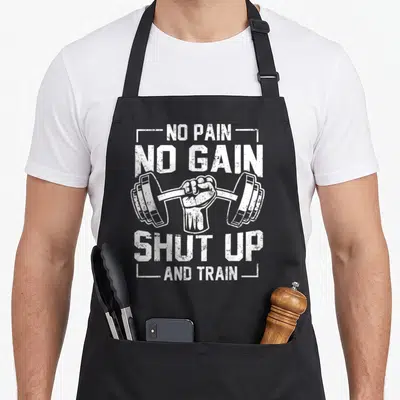 Tablier de cuisine noir avec poing serré tenant une barre de poids et texte blanc humoristique sur le sport.