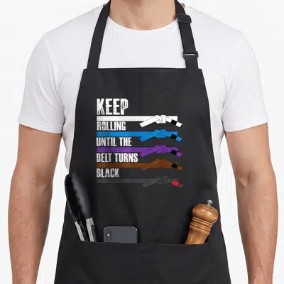 Tablier de cuisine noir avec cinq ceintures de jiu-jitsu et le texte Keep Rolling Until the Belt Turns Black imprimé en blanc.