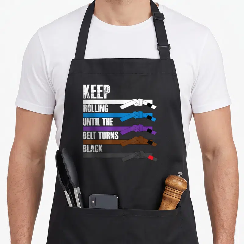 Tablier de cuisine noir avec cinq ceintures de jiu-jitsu et le texte Keep Rolling Until the Belt Turns Black imprimé en blanc.