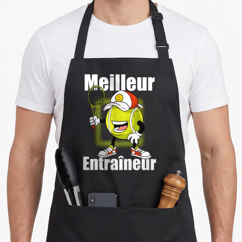 Tablier de cuisine noir avec un ballon de tennis anthropomorphique souriant tenant une raquette et le texte Meilleur Entraîneur.