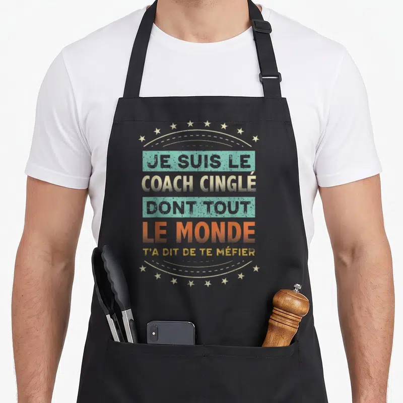 Tablier de cuisine noir avec inscription Je suis le coach cinglé dont tout le monde t'a dit de te méfier entouré d'étoiles dorées.