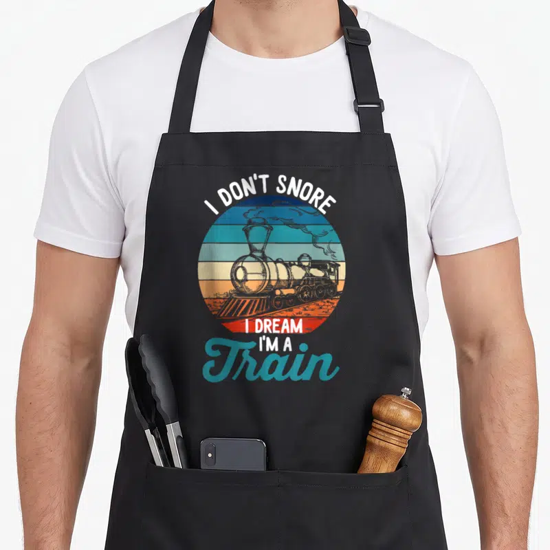 Tablier de cuisine noir avec une locomotive vintage et le texte I don't snore I dream I'm a Train sur un fond circulaire multicolore.