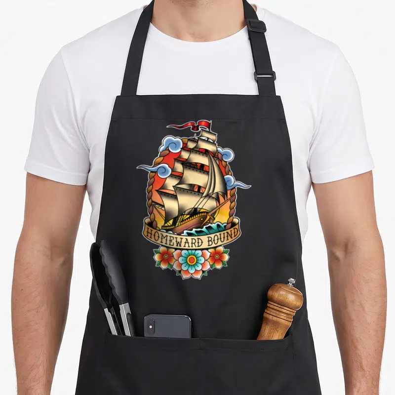 Tablier de cuisine noir avec illustration bateau à voile Homeward Bound style tatouage old school.