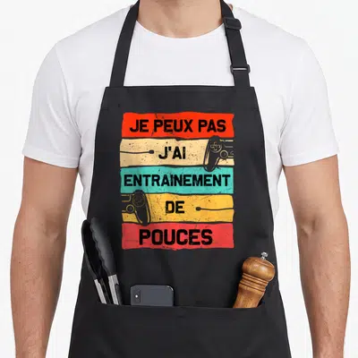 Tablier de cuisine noir avec bandeaux colorés et texte Je peux pas j'ai entrainement de pouces surmonté de manettes de jeu.