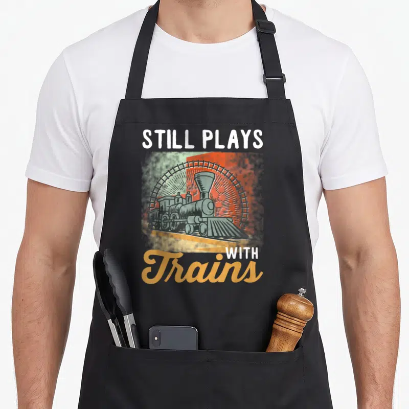 Tablier de cuisine noir avec illustration de locomotive vintage et texte Still Plays With Trains en blanc et orange.