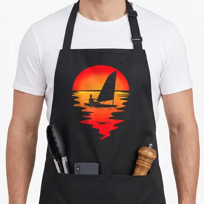 Tablier de cuisine noir avec illustration d'un bateau à voile devant un grand soleil couchant rouge et orange.