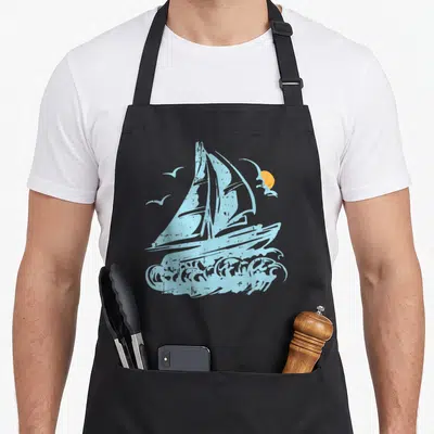 Tablier de cuisine noir avec un bateau à voile bleu, des oiseaux et un soleil orange sur fond gris uni.