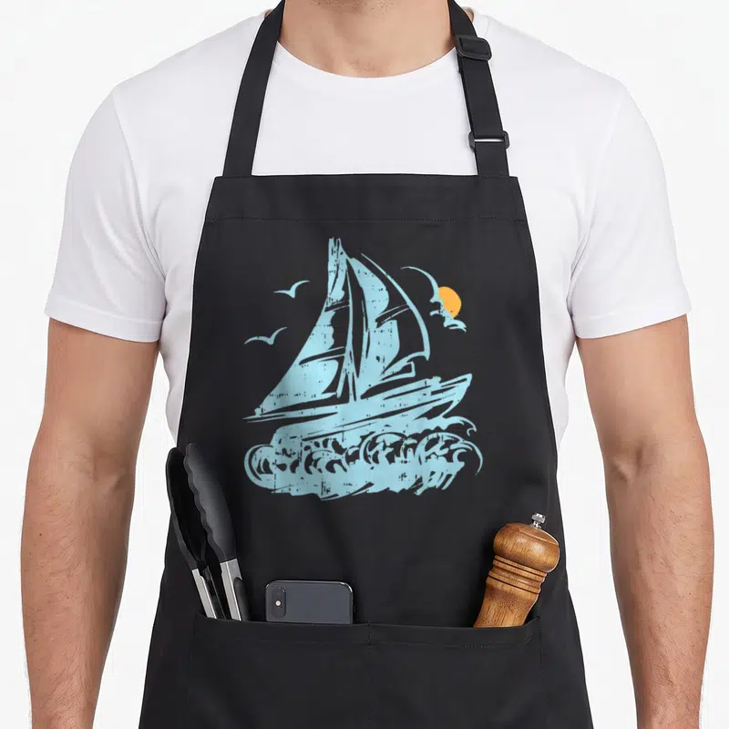 Tablier de cuisine noir avec un bateau à voile bleu, des oiseaux et un soleil orange sur fond gris uni.