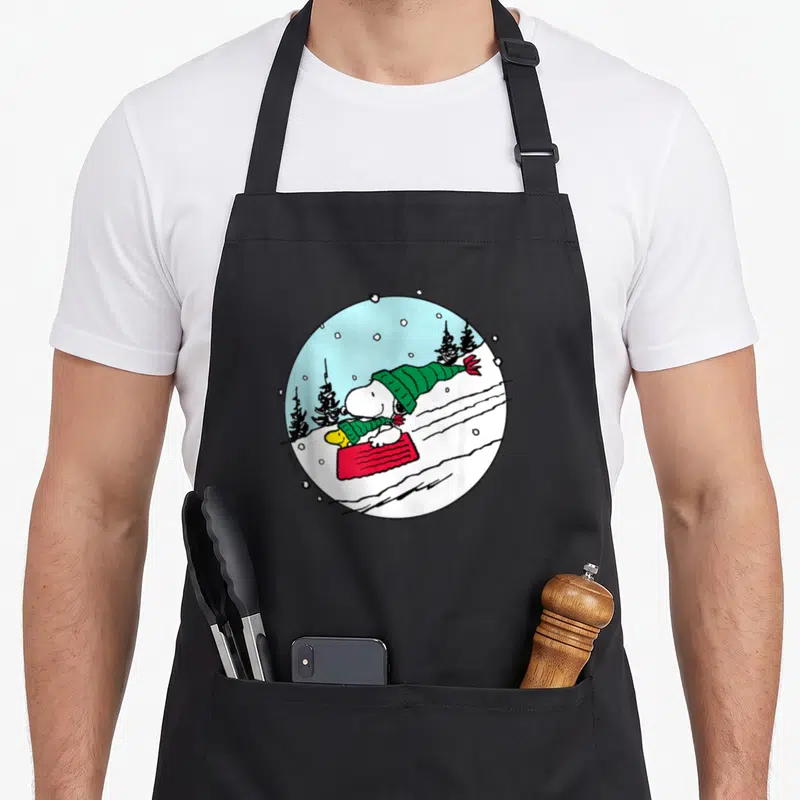 Tablier de cuisine noir avec illustration circulaire Snoopy traînant sur un patin rouge en compagnie de Woodstock dans la neige.