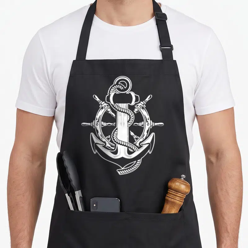 Tablier de cuisine noir avec ancre blanche enroulée de corde devant une barre de navire.