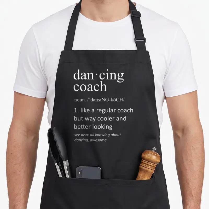 Tablier de cuisine noir avec l'inscription blanche Dan·cing coach et sa définition humoristique en anglais.