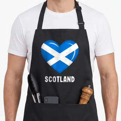 Tablier de cuisine noir avec un grand cœur stylisé aux couleurs du drapeau écossais et le texte Scotland en dessous.