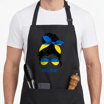 Tablier de cuisine noir avec une illustration de femme aux lunettes et bandeau aux couleurs du drapeau ukrainien.