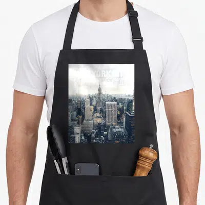 Tablier de cuisine noir avec une impression photographique du skyline de New York City et du texte blanc répété en surimpression.
