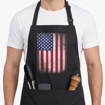 Tablier de cuisine noir avec le drapeau américain aux couleurs délavées et un aspect vintage usé sur toute la surface.