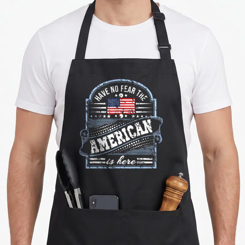 Tablier de cuisine noir avec un motif rectangulaire bleu clair contenant un drapeau américain et du texte en anglais blanc.