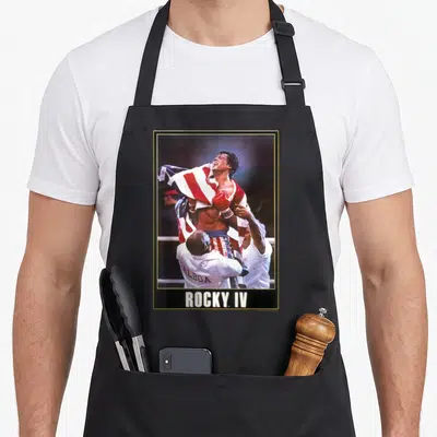 Tablier de cuisine noir avec une illustration de Rocky IV sous un drapeau américain et le texte ROCKY IV en blanc.