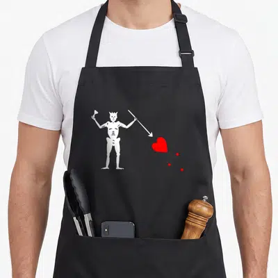 Tablier de cuisine noir avec illustration blanche d'un diable pointant une flèche vers un cœur rouge stylisé.