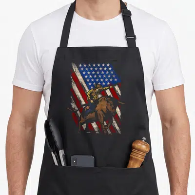 Tablier de cuisine noir avec illustration d'un cowboy sur un taureau devant un drapeau américain stylisé.