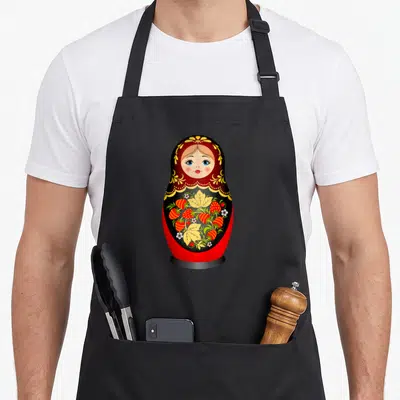Tablier de cuisine noir avec une poupée Matriochka rouge, noire et jaune décorée de fraises et de fleurs blanches.