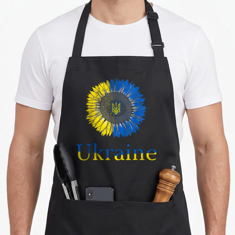 Tablier de cuisine noir avec un grand tournesol bicolore et le mot Ukraine imprimé en dessous.