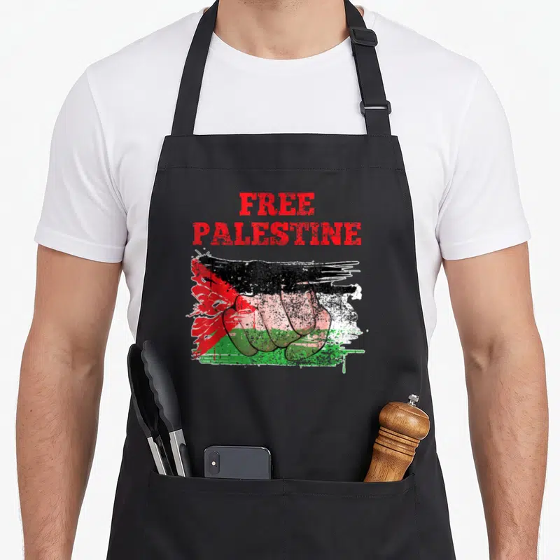 Tablier de cuisine noir avec un poing fermé coloré aux teintes du drapeau palestinien et du texte en rouge.