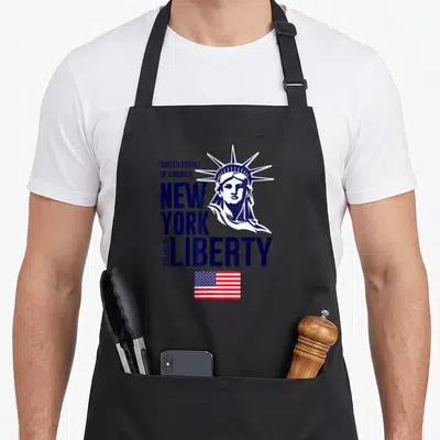 Tablier de cuisine noir avec illustration bleue et blanche de la Statue de la Liberté, texte New York et petit drapeau américain en bas.