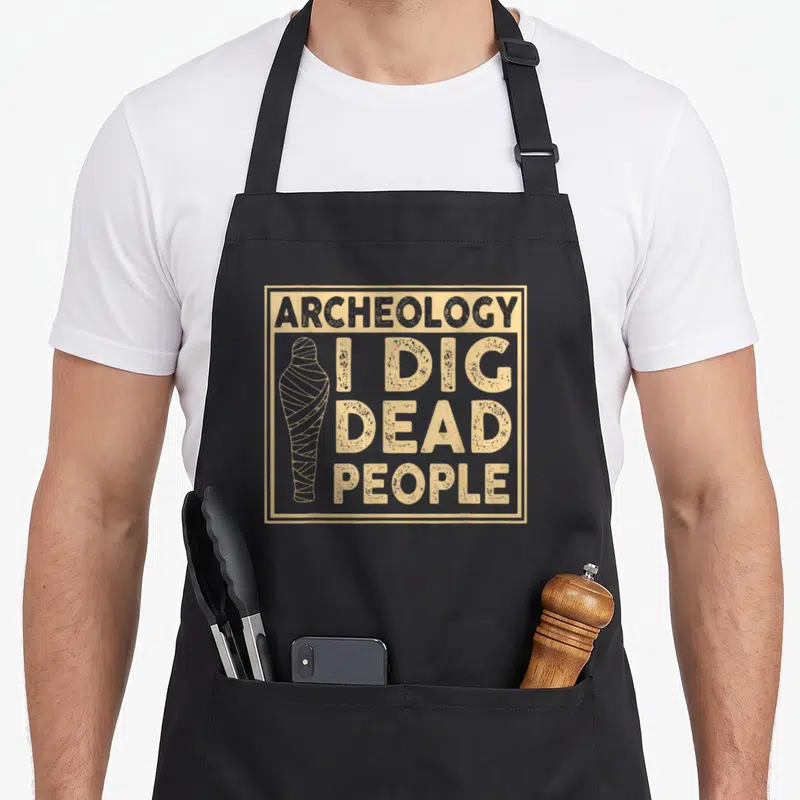 Tablier de cuisine noir avec inscription « I Dig Dead Gens » et dessin de momie beige.