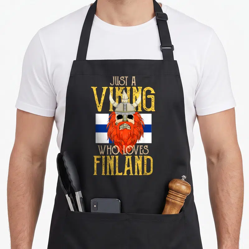 Tablier de cuisine noir avec un crâne de viking à barbe rouge sur fond de drapeau finlandais et texte jaune.
