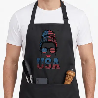 Tablier de cuisine noir avec illustration d'une femme au chignon et lunettes arborant les couleurs du drapeau américain sous l'inscription USA.