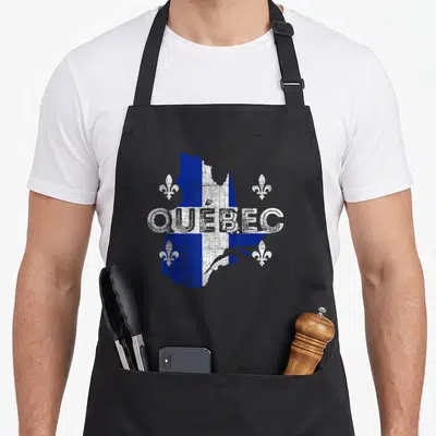 Tablier de cuisine noir avec carte du Québec en bleu et blanc, texte QUÉBEC et fleurs de lys blanches sur fond vieilli.